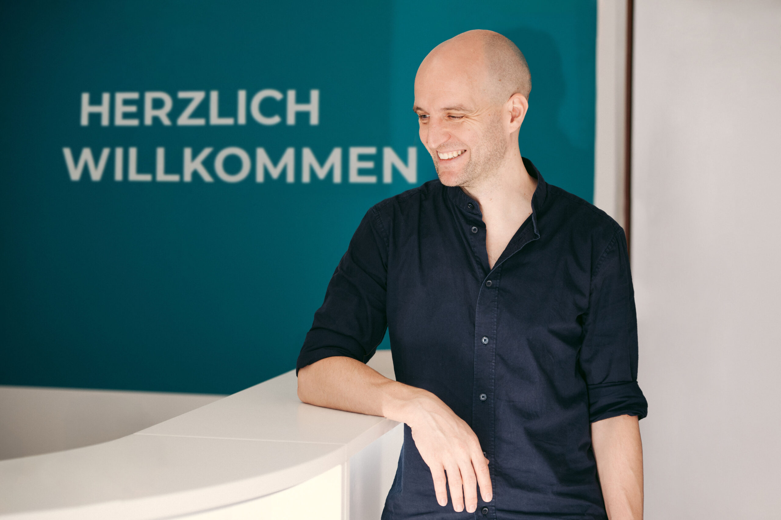 Philipp Gehrke blickt lächelnd zur Seite, hinter ihm der Willkommensschriftzug der Praxis.